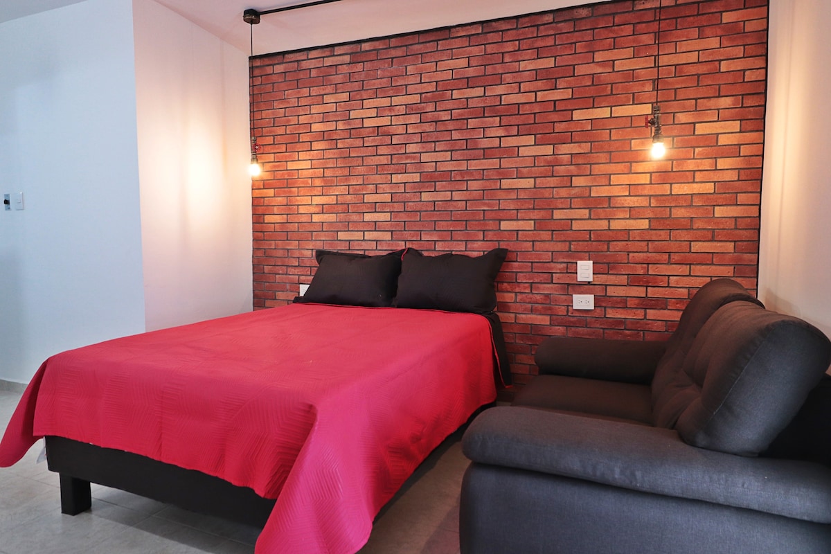 A cozy sleeping area features a spacious bed with a red blanket and dark pillows, complemented by a stylish gray sofa. The brick accent wall is highlighted by pendant lighting, creating a modern and inviting atmosphere.