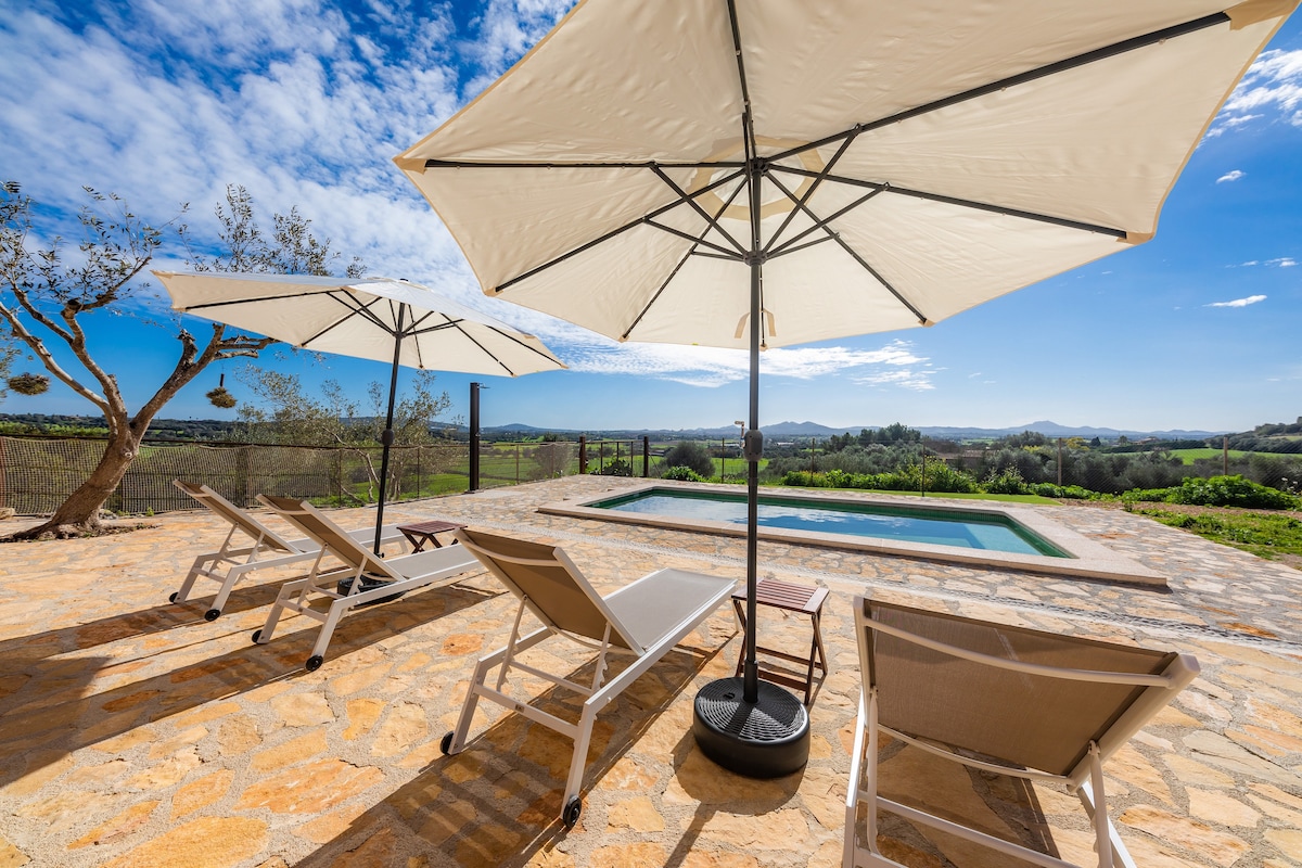 An outdoor area features a stone terrace with sun loungers and large umbrellas, providing shade. The inviting pool lies nearby, framed by views of the rolling landscape and mountains. Clear skies and natural light enhance the serene setting.