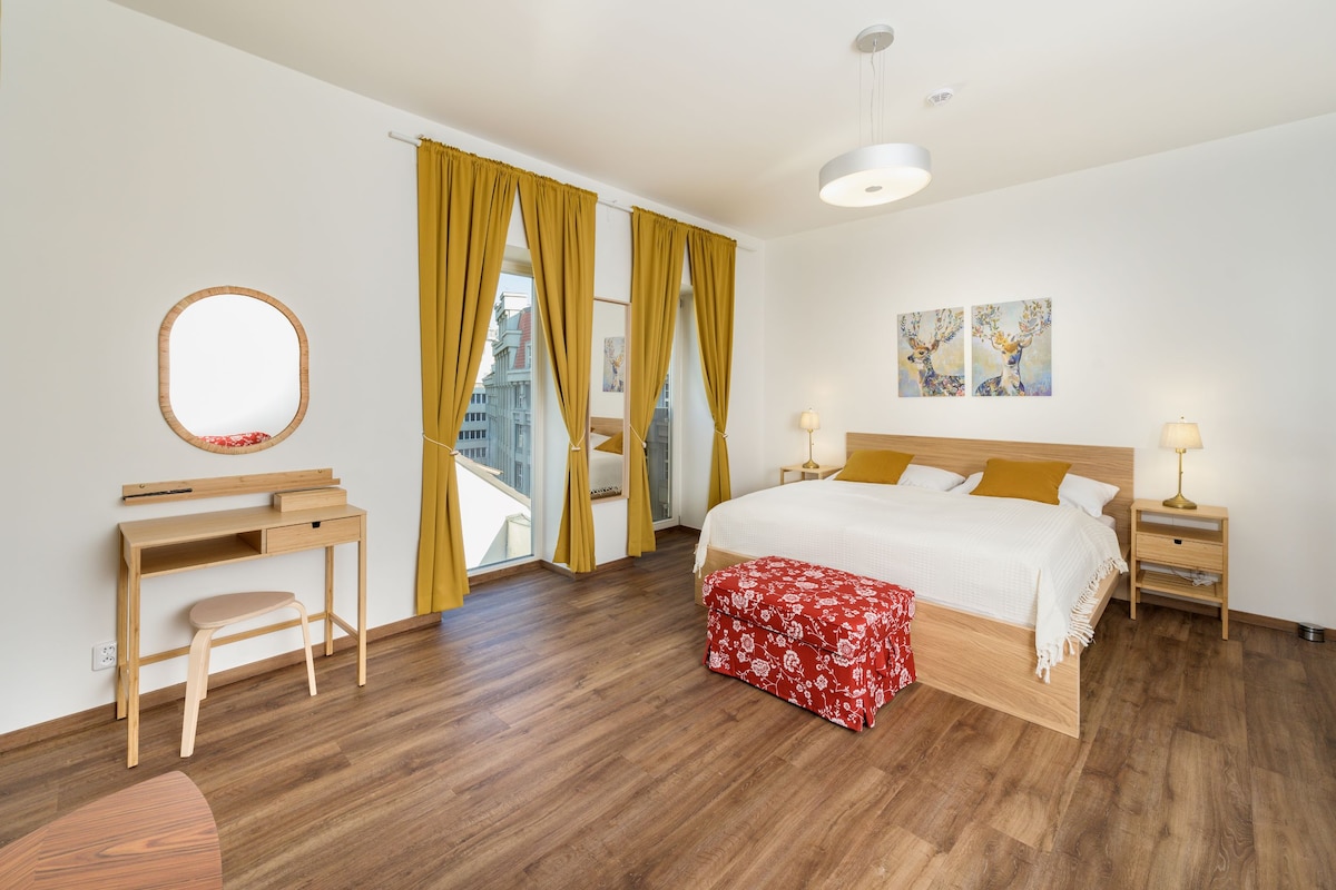 A spacious bedroom is illuminated by natural light through large windows with yellow curtains. A large bed with white linens is positioned against the wall, accompanied by bedside tables with lamps. A round mirror and a small desk are featured, creating a functional and inviting space.