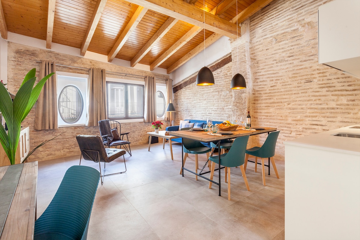 A spacious living and dining area is highlighted by a wooden beam ceiling and exposed brick walls. The room features a comfortable sofa, a dining table seating four, and stylish chairs. Bright natural light enters through two large windows, enhancing the welcoming ambiance.