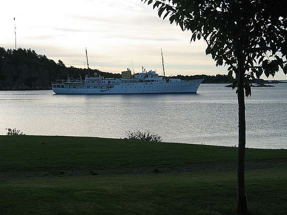 A large white ship is anchored in calm waters, displaying its smooth hull and several masts. The background features a green landscape with trees, and the water reflects the soft colors of the sky. Gentle ripples can be seen on the surface.