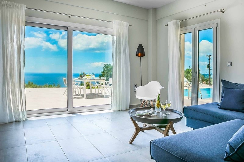 A bright living area is presented with large glass doors allowing panoramic views of the sea and outdoor dining space. A light-colored sofa sits beside a wooden coffee table, with refreshing drinks set on it. Sheer curtains frame the glass doors, enhancing the open feel of the space.