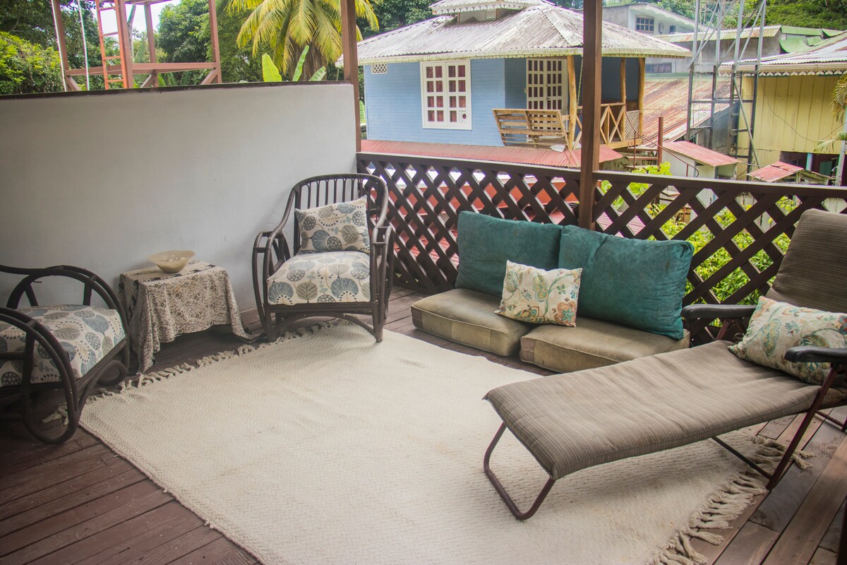 A shaded terrace features comfortable seating with patterned cushions, including a spacious sofa and two lounge chairs. A neutral area rug covers the wooden floor, with a small table positioned in the corner. Lush greenery and nearby colorful houses can be seen in the background.