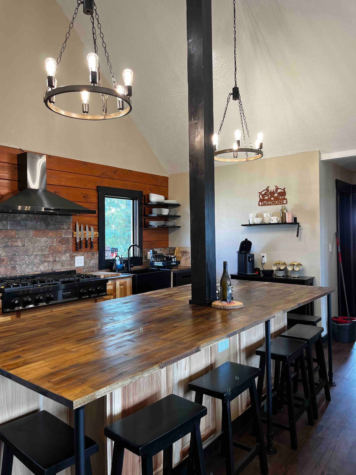 A spacious chef’s kitchen features a large wooden counter with black stools. Two stylish pendant lights hang from the high ceiling above. Equipped with a professional gas stove and ample cabinetry, the kitchen offers a warm and functional space for meal preparation.