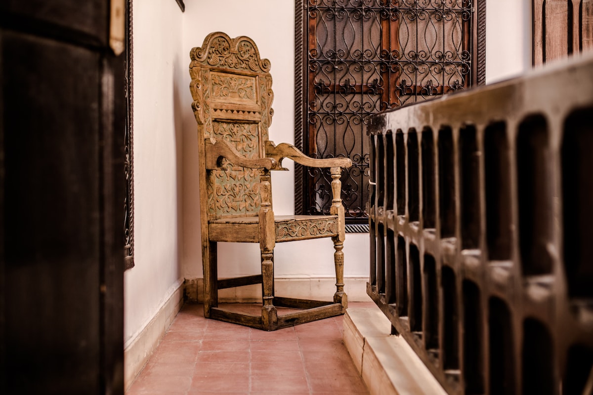 An intricately carved wooden chair occupies a corner of a corridor. The chair features a high back and armrests, showcasing detailed craftsmanship. Surrounding it are traditional ironwork details and patterned tiles, enhancing the space's cultural ambiance.
