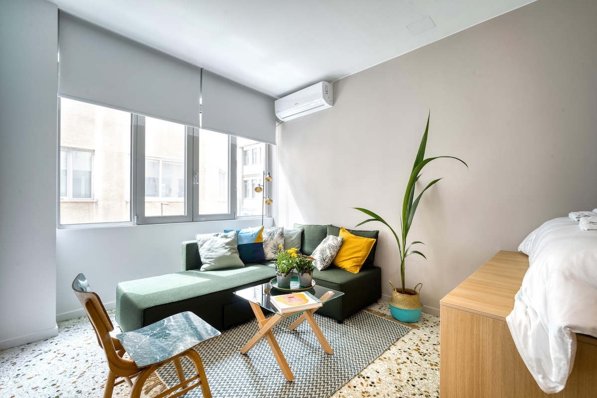 A bright living area features a green sectional sofa adorned with colorful pillows. A wooden coffee table sits on a classic mosaic floor, surrounded by complementary decor. Large windows allow natural light to fill the space, while a potted plant adds a touch of greenery.