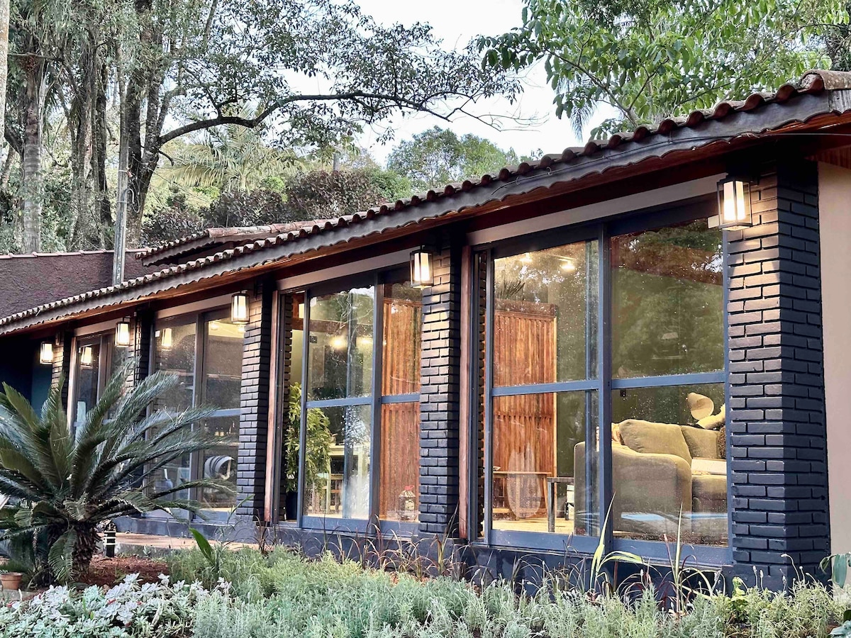 A single-story building is shown with large windows framed in dark tones, allowing natural light to illuminate the interior. Lush greenery surrounds the structure, including carefully arranged plants and a landscaped garden, contributing to a serene outdoor ambiance.
