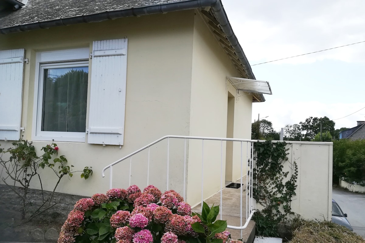 A light-colored exterior of the house is shown, with a set of white shutters framing a large window. Steps lead up to the entrance, surrounded by vibrant flowering plants, adding a touch of greenery to the scene.