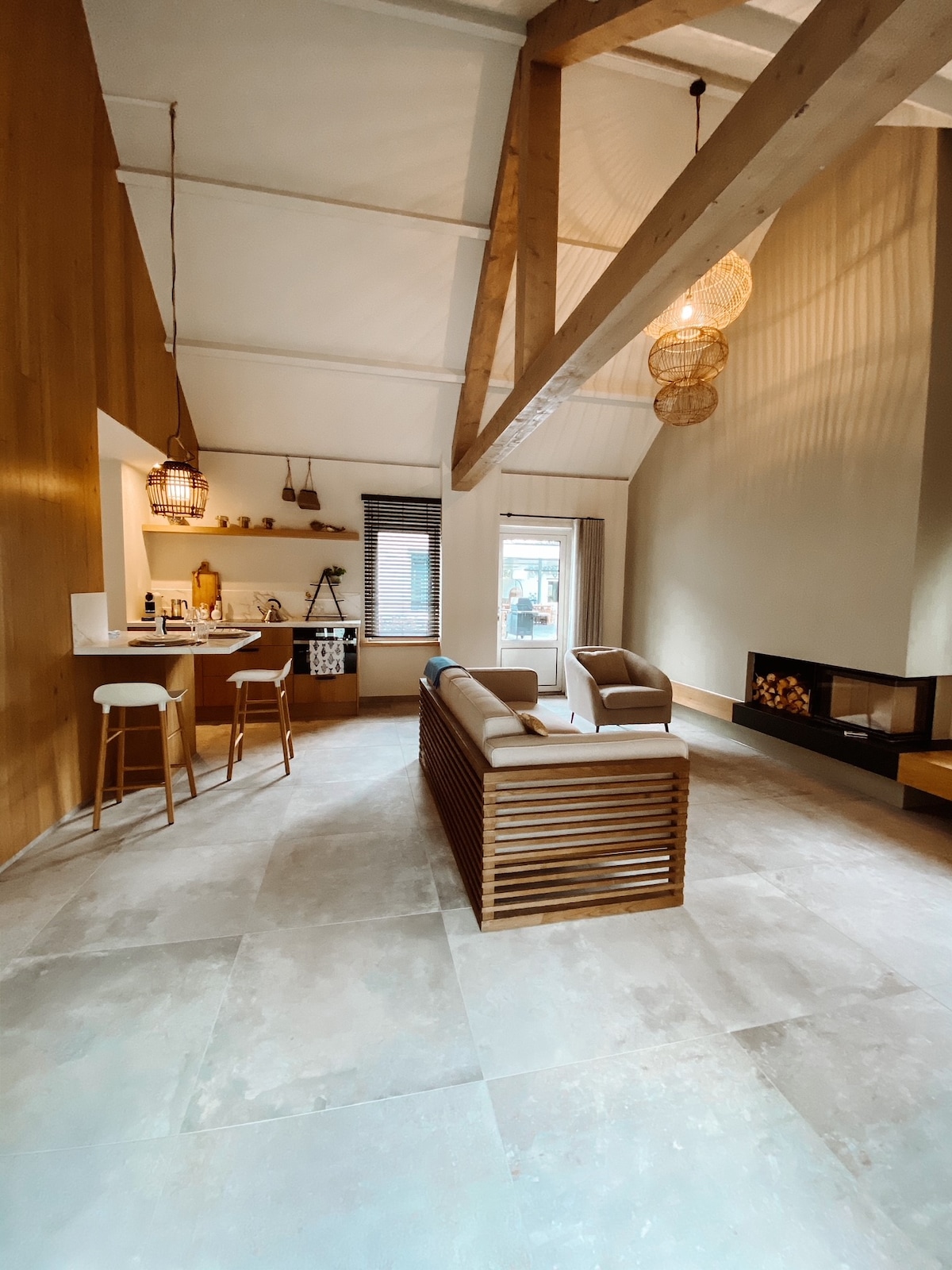 An open living area features a modern kitchen with high stools, complemented by wood accents and decorative lanterns. A spacious seating area includes a light-toned couch and armchairs, while a sleek fireplace adds warmth to the space. Natural light fills the room through large windows.