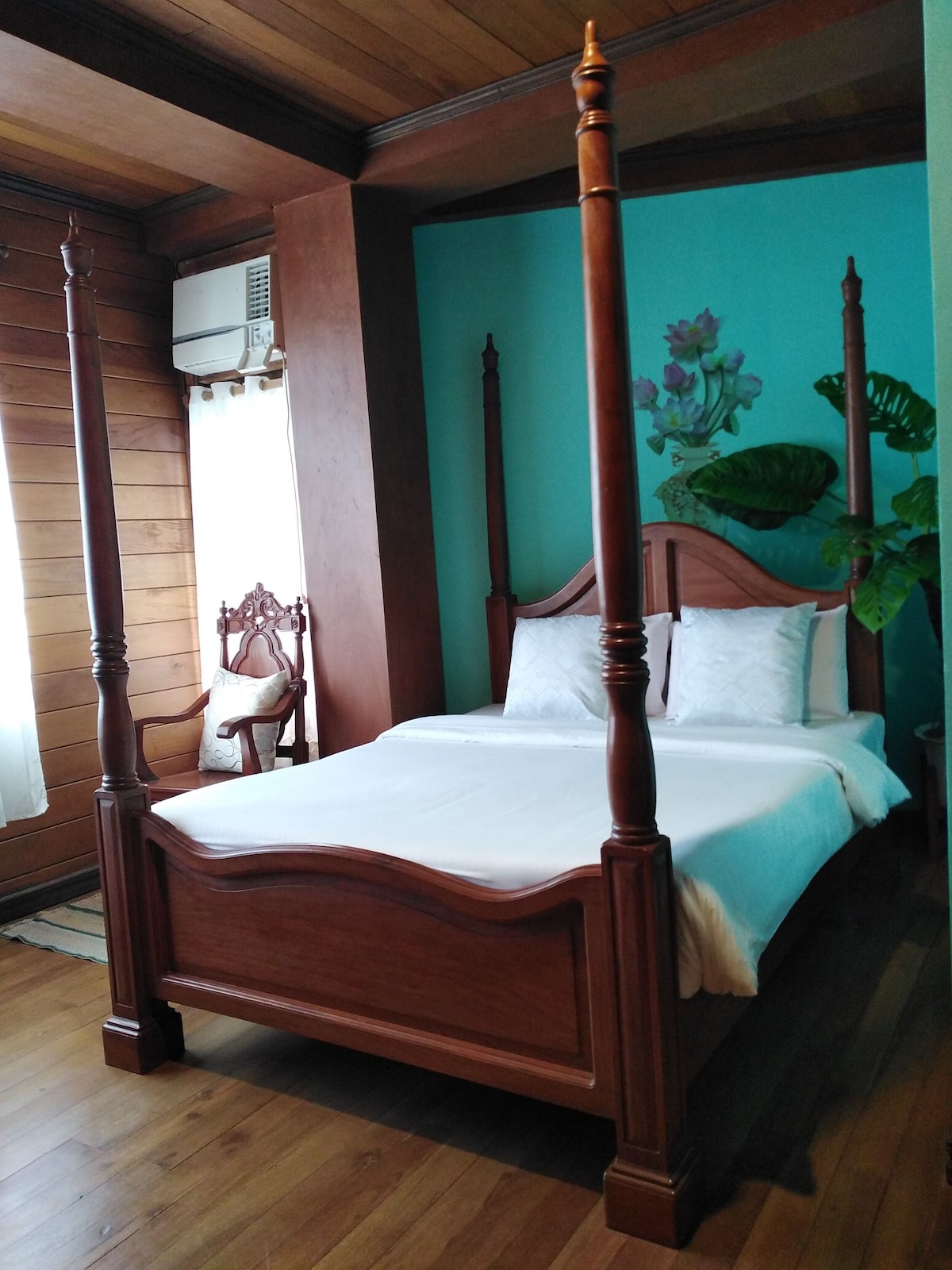 A spacious bedroom features a wooden four-poster bed with a crisp white linen arrangement. A vibrant turquoise wall is complemented by natural wood accents. A chair sits in one corner, and light enters through a nearby window, providing a bright atmosphere.