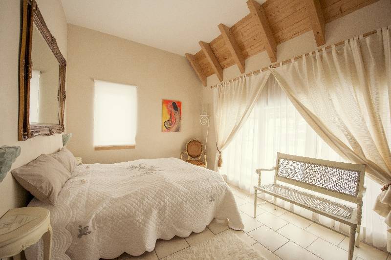 A spacious bedroom features a large double bed with a textured white quilt. Natural light filters through sheer curtains, illuminating the warm tones of the wood-beamed ceiling. A vintage bench and a round mirror add character to the inviting space.