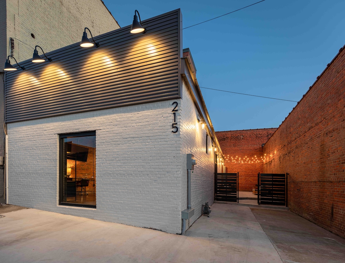The exterior of the studio apartment is illuminated by overhead lights, highlighting the sleek design of the building. A large window offers a view into the interior, while a well-maintained outdoor area is visible beside the structure, bordered by brick walls.