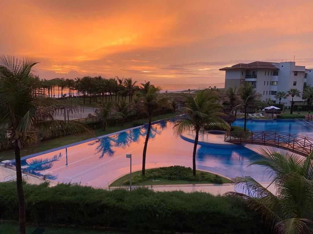 A serene view features a large swimming pool, reflecting hues of orange and purple from the sunset. Lush palm trees surround the area, while a modern building is visible in the background, offering a tranquil setting near the beach.