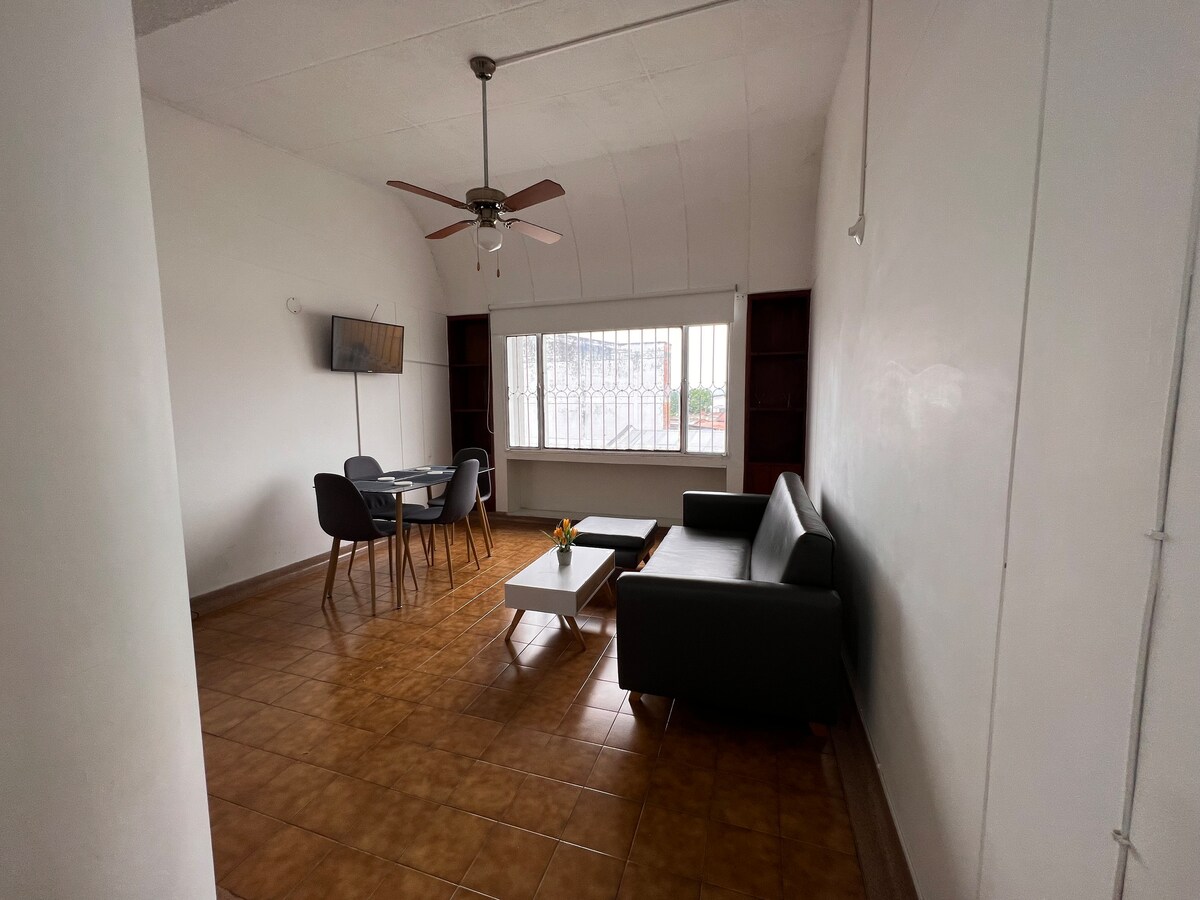 An open living area features a light-colored sofa, a modern coffee table, and a small dining table with chairs. A ceiling fan hangs above, while a window allows natural light to fill the space. A wall-mounted TV is positioned nearby.