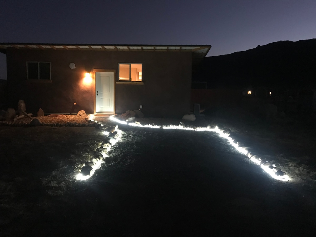 The exterior of the casita is illuminated by soft lighting along the path leading to the entrance. A large window reflects the warm glow of indoor lights, while the surrounding landscape hints at the natural beauty of the area under a twilight sky.