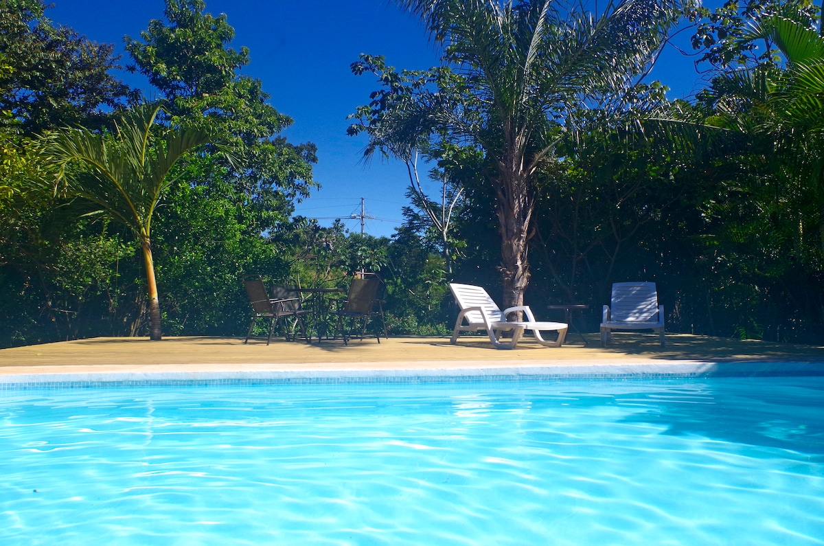 A serene pool area is framed by lush greenery, featuring clear blue water reflecting the sky. Lounge chairs are positioned beside the pool, alongside a small outdoor dining set, creating a relaxing space for guests to enjoy.