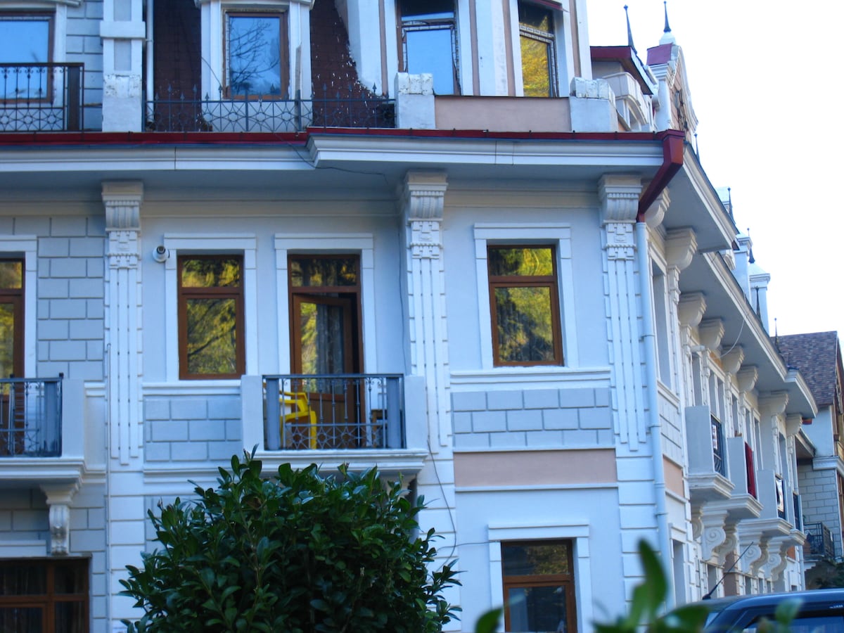 The exterior of a charming multi-story building is visible, showcasing decorative architectural features. Balconies with wrought-iron railings are situated above large windows, reflecting the surrounding greenery. A lush hedge is seen in the foreground, adding to the inviting appeal of the entrance.