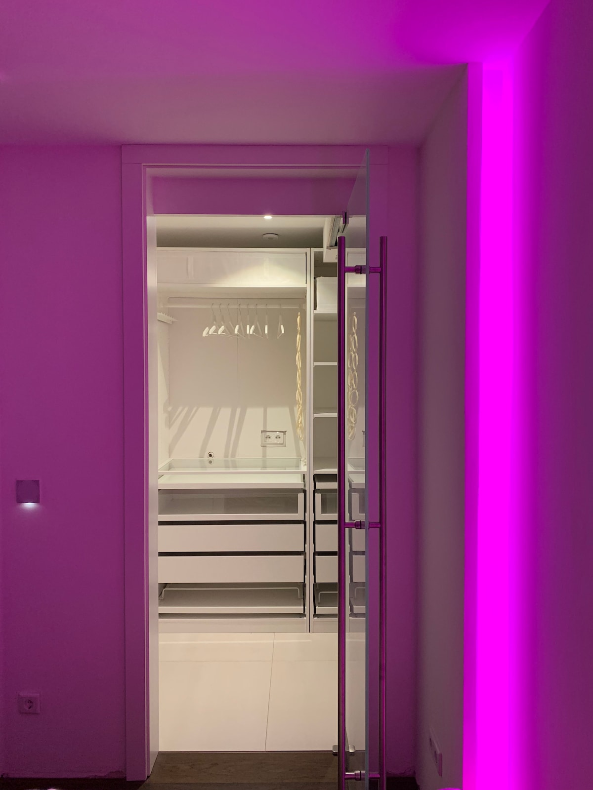 A well-organized walk-in closet is visible through a glass door, featuring built-in shelves and drawers for storage. Soft lighting enhances the space, while a vibrant pink accent light adds a modern touch to the surroundings.
