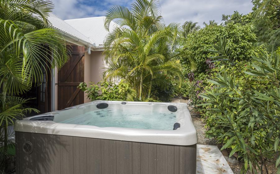A private hot tub is surrounded by lush greenery, featuring tropical plants and palm trees. The structure is situated along a pathway, providing a serene escape in a natural setting. Soft sunlight filters through the leaves, creating a tranquil atmosphere.