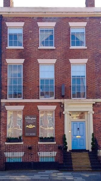 A three-story brick townhouse features large windows with white trim and decorative curtains. The entrance is highlighted by a blue door and flanked by symmetrical potted plants. A sign indicating the Georgian Townhouse Hotel is displayed prominently on the facade.
