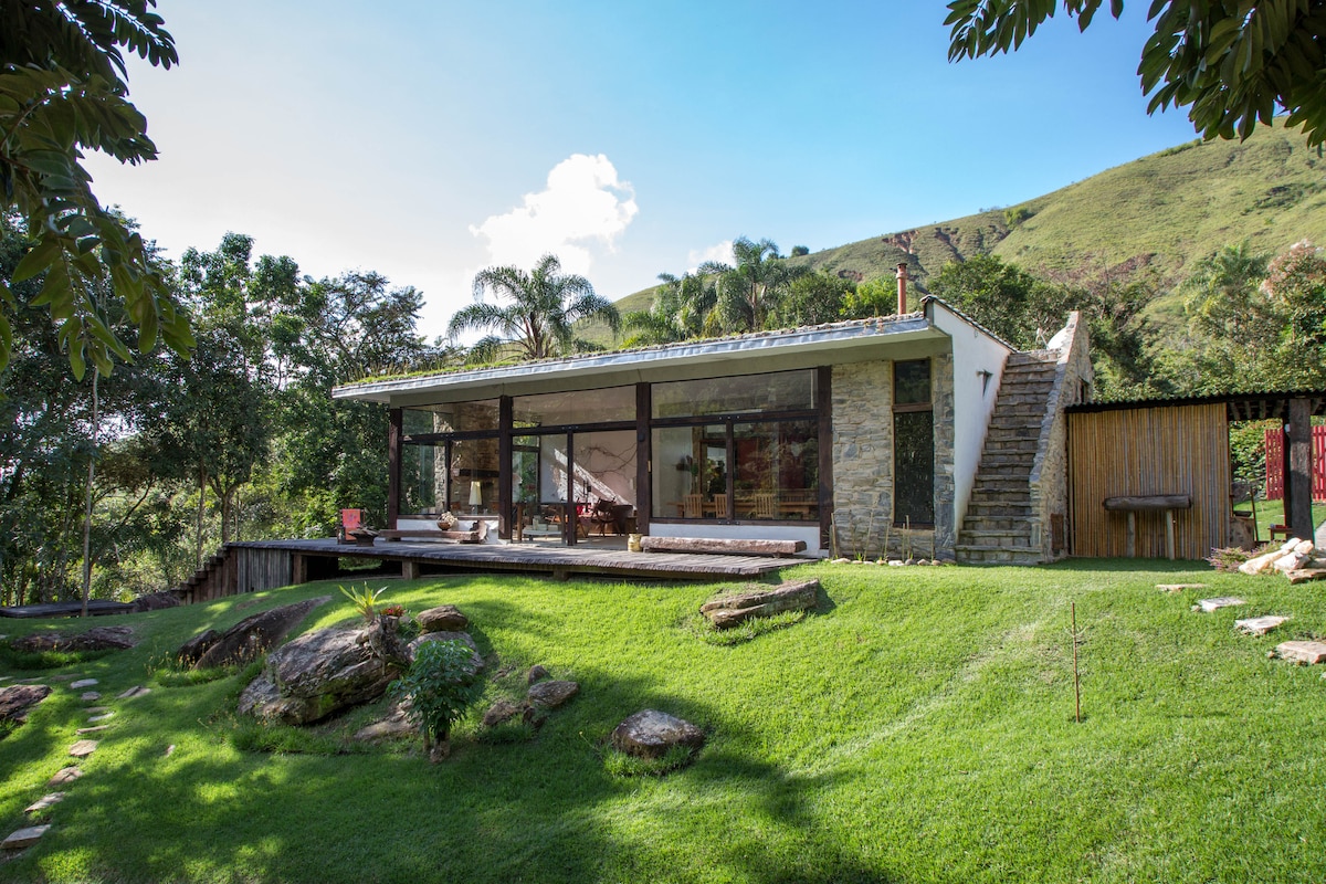 A modern house with a stone and glass exterior is situated on a lush green lawn. A spacious deck extends out, surrounded by native plants. The backdrop features rolling hills and a clear blue sky, creating a serene setting for relaxation.