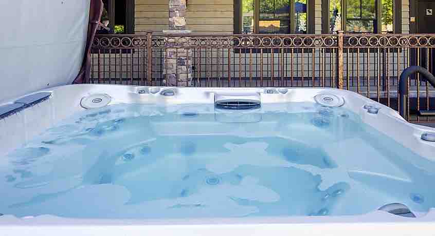 A jetted hot tub is positioned in a private area, displaying clear water with visible jets along the sides. Surrounding fencing enhances the secluded ambiance, while the structure's exterior and windows are partially visible in the background.