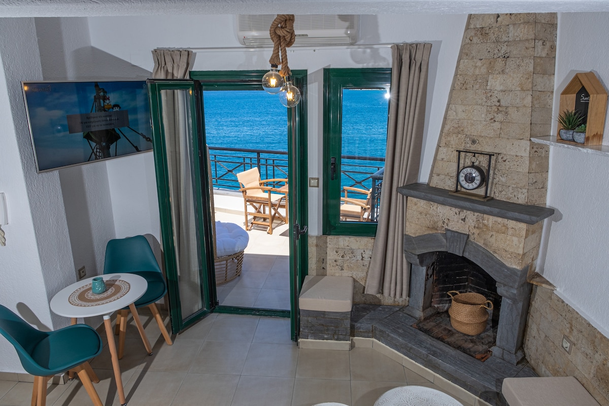 The living space is equipped with a cozy seating area featuring a fireplace. Large glass doors open onto a terrace with sea views. Natural light fills the room, complemented by decor that includes a flat-screen TV and light-colored furnishings.