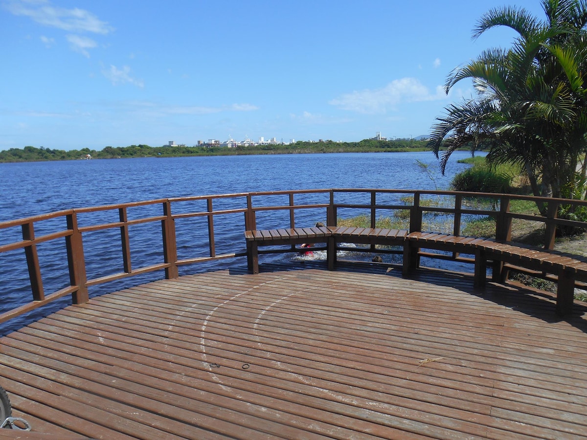 A wooden deck extends over a tranquil lagoon, providing a scenic view of water and greenery. Surrounding vegetation includes palm trees, enhancing the natural setting. The distance showcases a glimpse of town buildings against the clear blue sky.