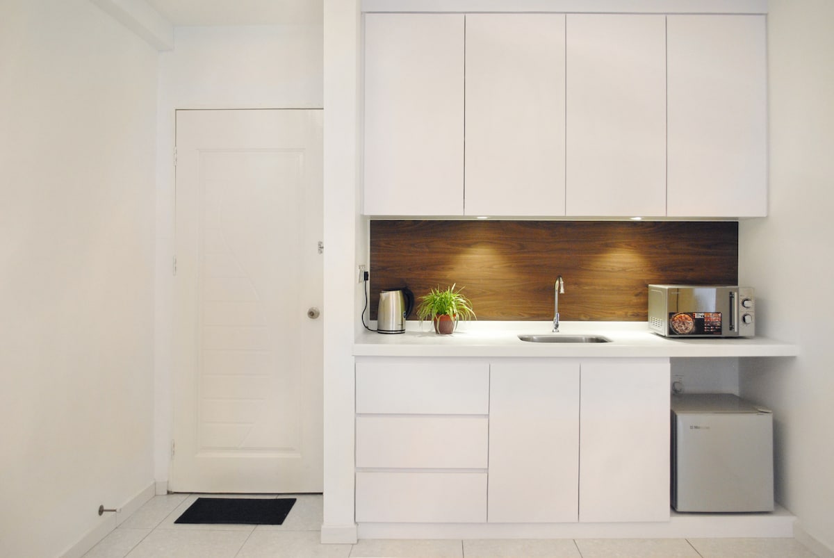 A modern kitchenette is featured, showcasing sleek white cabinets with a wooden backsplash. A small sink is positioned beneath overhead lighting, while a bar-sized refrigerator and microwave are visible. A kettle and a small plant add subtle accents to the space, enhancing functionality.