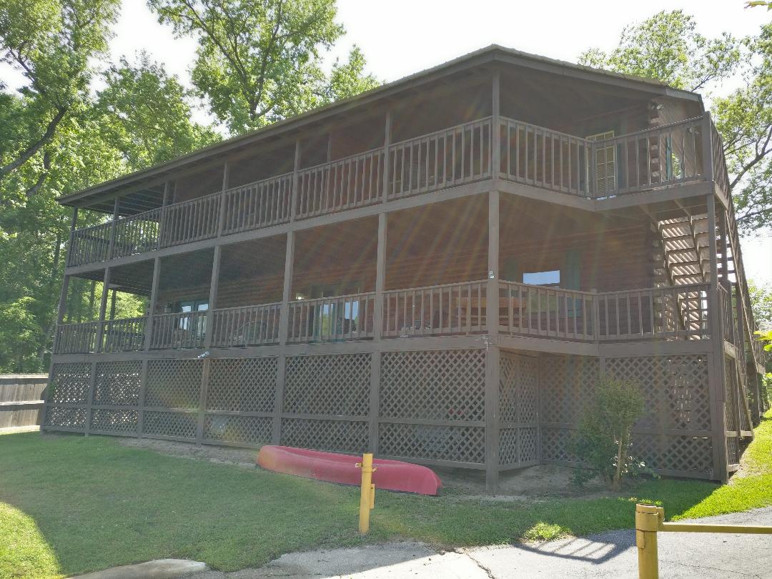 A three-story wooden structure is set amidst greenery, featuring multiple balconies and expansive deck space. The exterior showcases a natural wood finish with a light brown hue. A kayak rests on the grass, indicating access to water activities nearby.