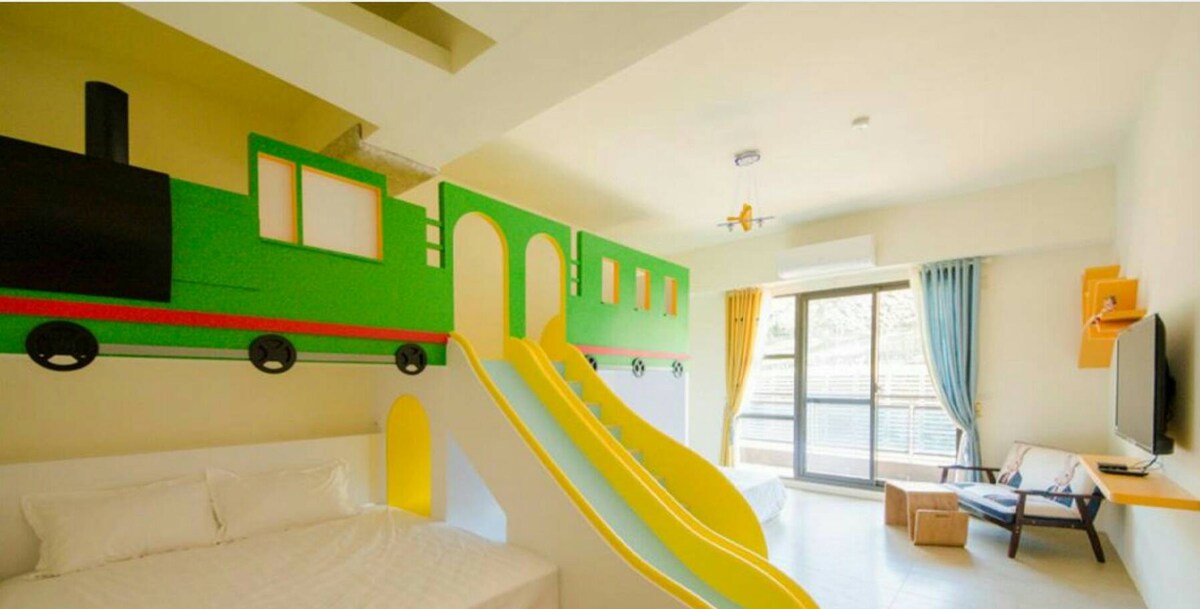 A playful room features a vibrant train-themed wall design, complemented by a yellow slide. A spacious bed is positioned at the side, with natural light filtering through large windows adorned with light curtains. A media unit is visible across from the seating area.