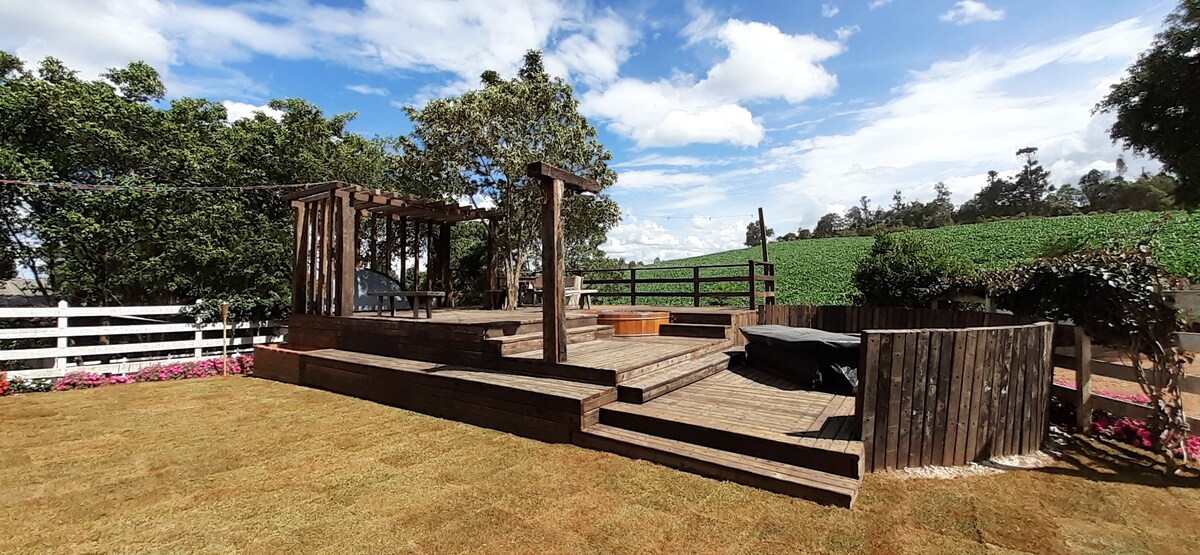 A wooden deck area is presented, complemented by a hot tub filled with water. Surrounded by trees and grassy space, the deck includes a pergola structure. A picturesque view of the surrounding fields is visible in the background, enhancing the outdoor setting.