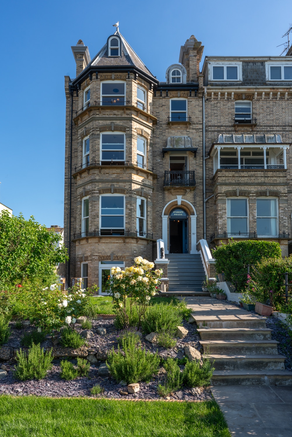 The Victorian-style building features a three-story façade adorned with ornate architectural details. A neatly landscaped garden with green grass and flowering plants surrounds the entrance, which is reached by a stone walkway and a set of elegant stairs leading to a welcoming front door.