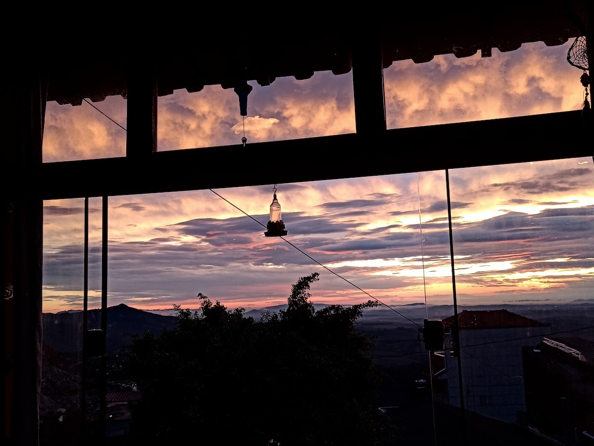 A stunning view is captured through large windows, showcasing a vibrant sunset with swirling hues of orange and purple. Silhouettes of distant mountains create a serene backdrop, with a decorative light fixture hanging above the scene, enhancing the ambiance of the space.