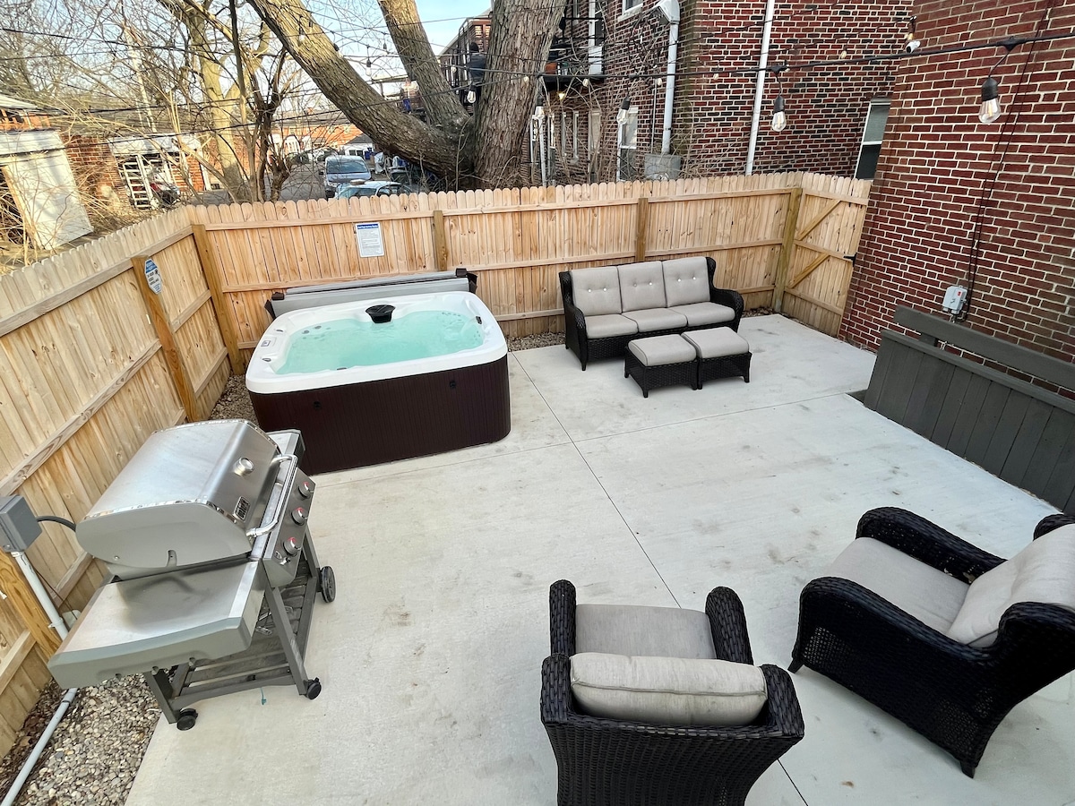 An outdoor space is visible, featuring a built-in hot tub surrounded by a concrete patio. Comfortable seating arrangements include a gray sectional sofa and two chairs. A grill is positioned nearby, with a wooden fence enclosing the area for privacy.