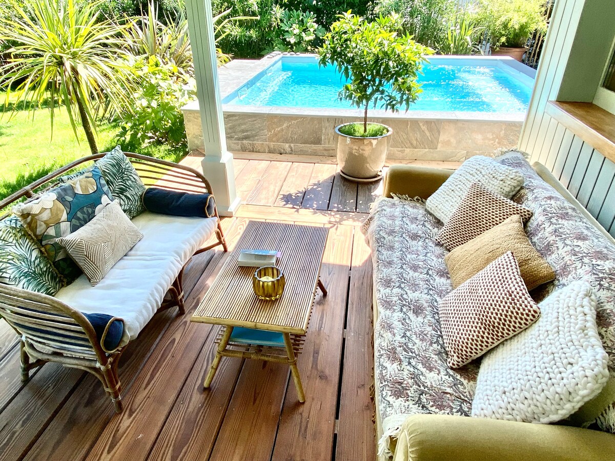 A covered outdoor seating area features comfortable couches adorned with soft cushions, inviting relaxation. A small table sits in the center, with a decorative bowl resting atop. In the background, a private pool reflects the surrounding greenery, enhancing the tranquil atmosphere.
