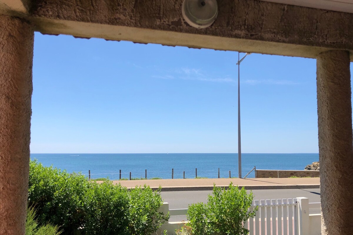 A view of the ocean is framed by two stone pillars, showcasing a clear blue sky and calm waters. The horizon is visible, along with the road and a white fence in the foreground, enhancing the coastal setting.