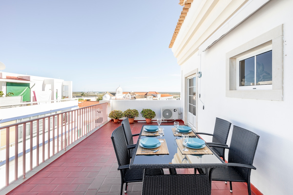 An outdoor dining area is visible on the balcony, featuring a table set for four with blue plates and woven placemats. Surrounding potted plants add a touch of greenery, while the distant skyline highlights Faro's architecture and natural beauty.