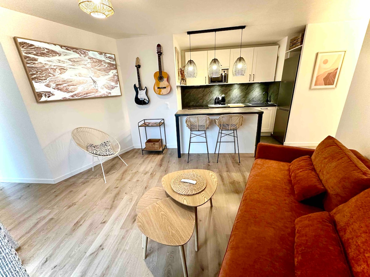 An inviting living space features a cozy orange sofa and two wooden side tables. A stylish wall art piece adds a touch of character, while a guitar and an acoustic guitar are displayed, enhancing the aesthetic. A well-equipped kitchen is visible in the background, with modern fixtures and a green marble backsplash.