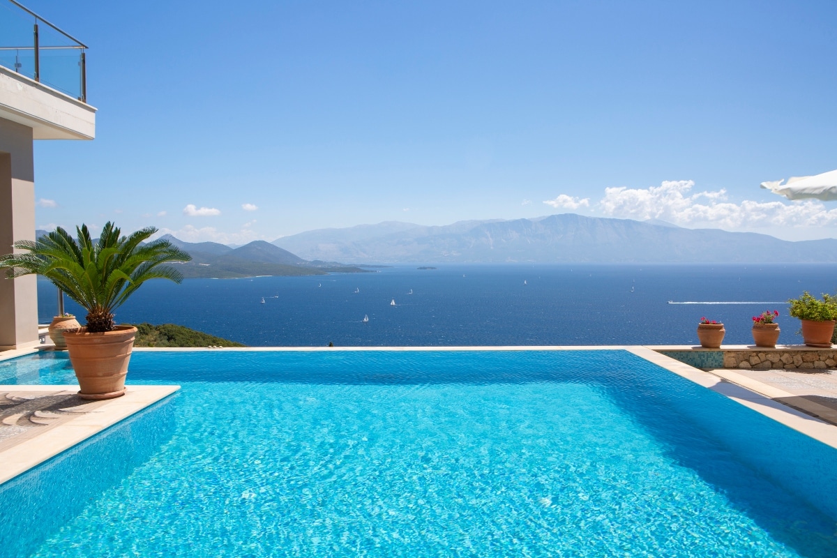 An infinity pool reflects the clear blue sky and the scenic Ionian Sea, surrounded by potted palm trees and vibrant flowers. The terrace features a spacious area for relaxation, overlooking the tranquil waters and distant mountains.
