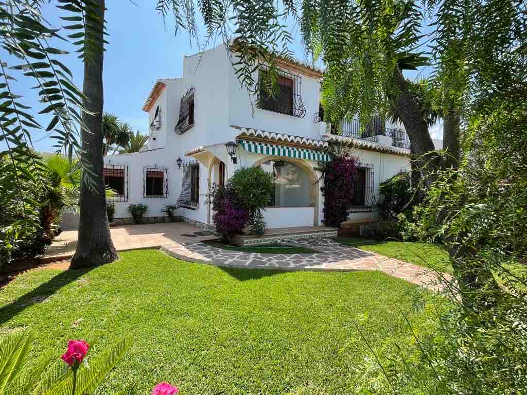 A white two-story building is framed by lush green grass and tropical plants. The facade features decorative shutters and vibrant flowers, creating an inviting entrance. A stone pathway leads to the front door, and trees provide shade in the surroundings.