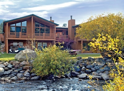 The exterior of a wooden lodge is depicted, featuring large windows and multiple balconies overlooking a gently flowing river. Nearby, rocks and lush greenery frame the scene, enhancing the connection to nature and the surrounding landscape.