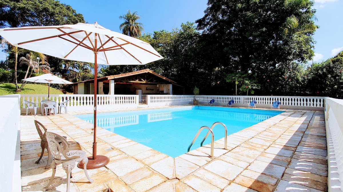 A clear blue swimming pool is framed by a tiled patio, featuring white umbrellas providing shade. Lounge chairs are positioned around the pool, with a wooden structure visible in the background, surrounded by greenery and palm trees under a bright sky.