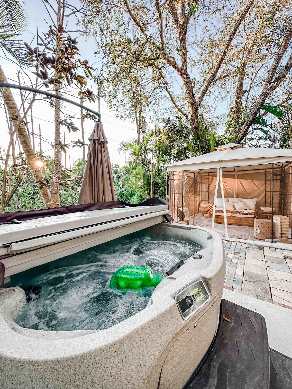 A serene hot tub is seen with bubbling water, situated amidst lush greenery. A shaded seating area featuring a tent with comfortable bedding is nearby, providing a peaceful retreat. The setting is enhanced by natural light filtering through the surrounding trees.
