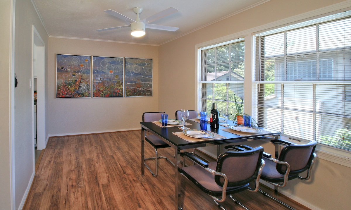 A dining area features a rectangular table set with plates and glasses, accommodating four seats. Colorful artwork adorns the wall, enhancing the space. Natural light filters through large windows, illuminating the hardwood floor and creating a cheerful ambiance.