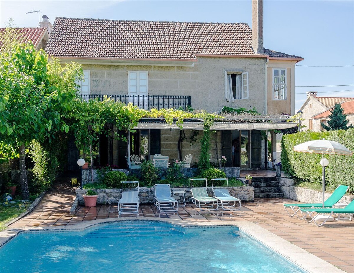 The exterior of a spacious house is visible, featuring a private pool surrounded by sun loungers. A shaded patio area houses comfortable seating, with greenery framing the space. Windows on the upper floor allow natural light to filter into the home.
