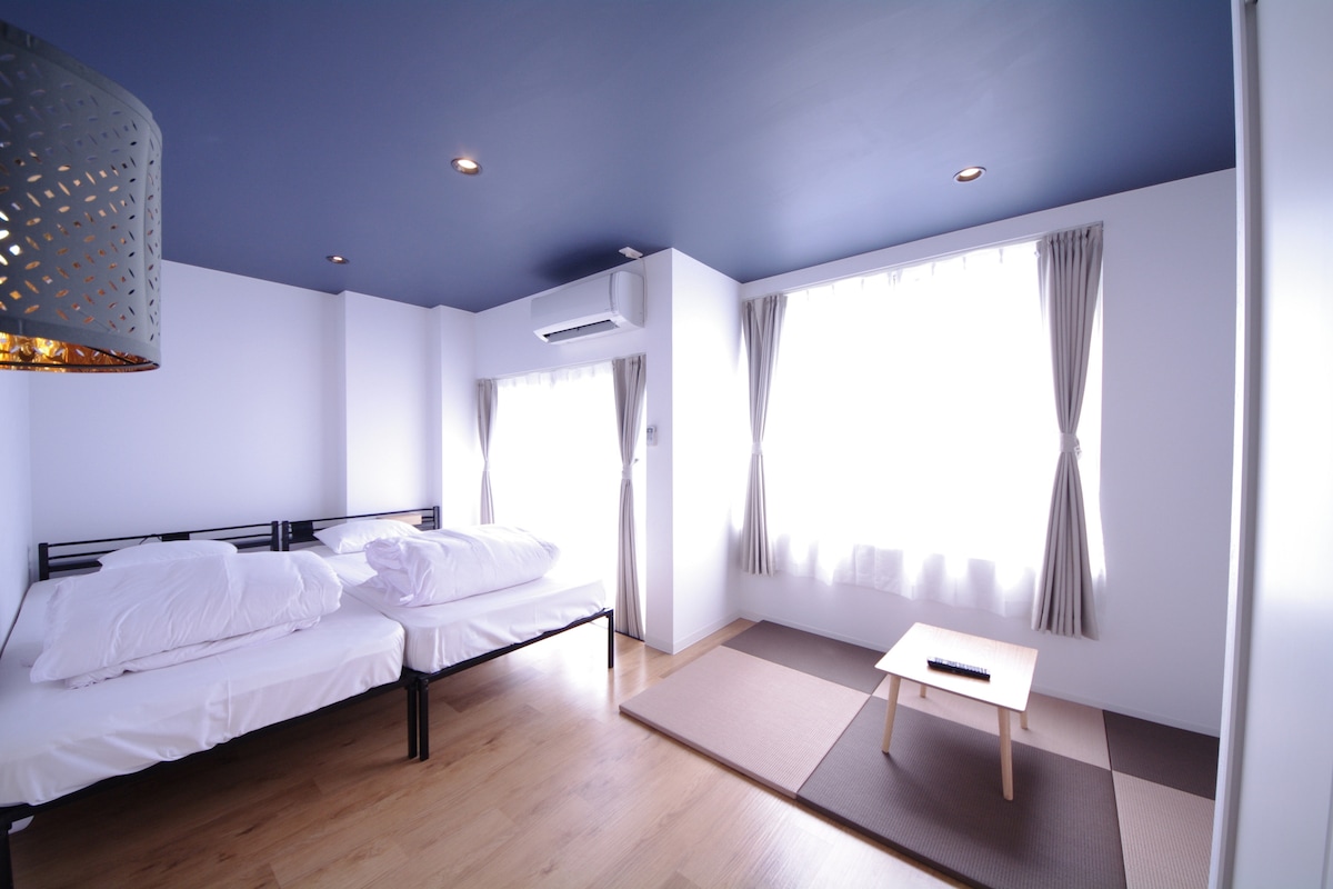 A room is presented with two spacious beds adorned with neatly arranged bedding. Beige tatami flooring contrasts with the light-colored walls. A low wooden table is situated on the tatami mat. Natural light filters through large windows covered with light curtains.