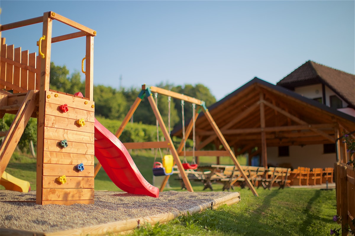 A wooden playground features a climbing structure, slide, and swings, set on a soft, gravel surface. In the background, a spacious covered terrace provides shaded seating for dining, with lush greenery and vineyards surrounding the area.