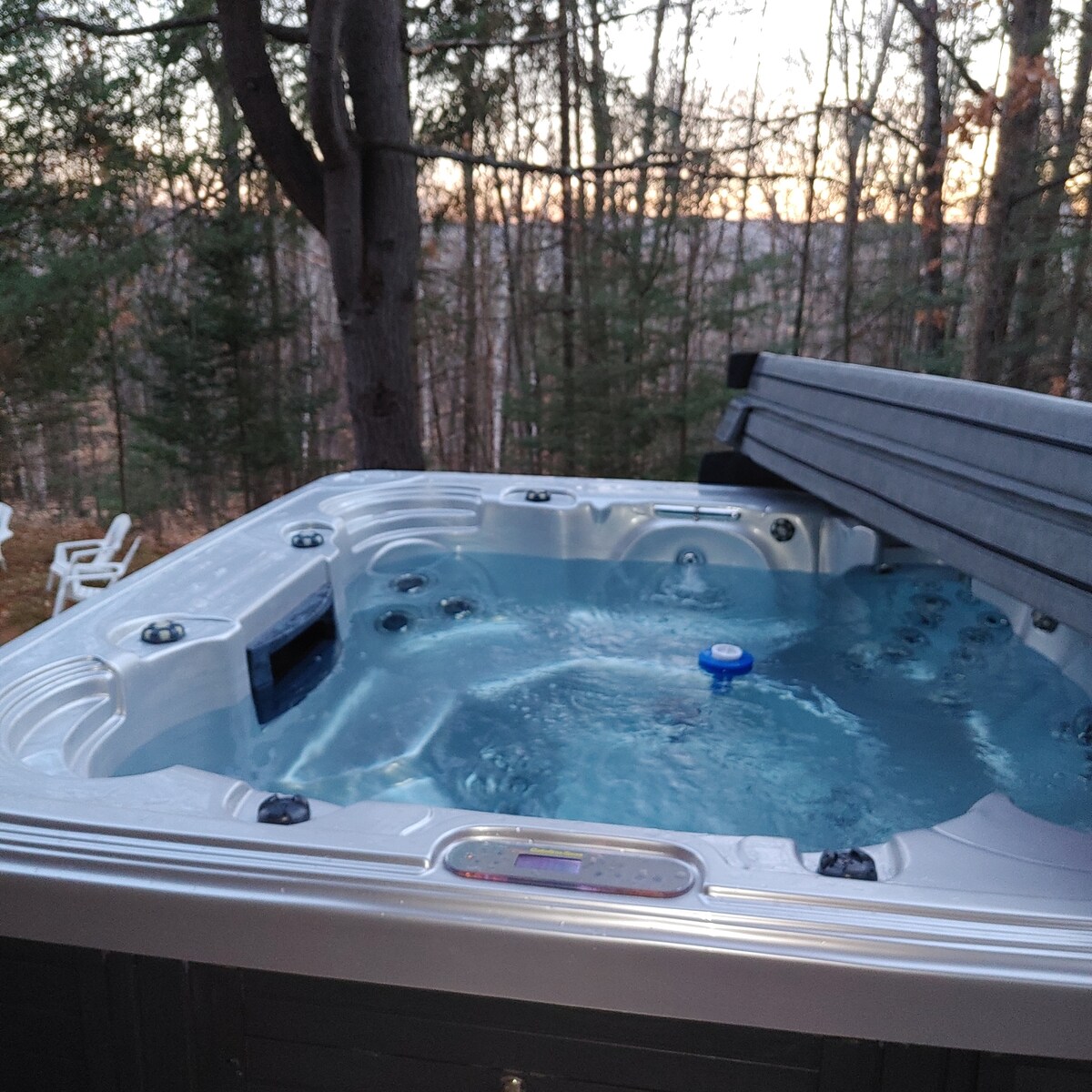 A hot tub is featured on a deck surrounded by trees, with water gently bubbling in the evening light. The setting offers a serene view of the forest, providing a tranquil atmosphere for relaxation.