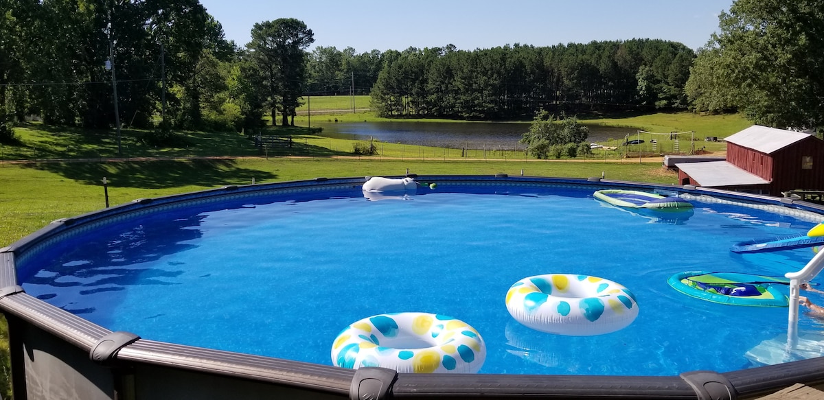 An above-ground swimming pool is surrounded by a spacious grassy area, featuring colorful inflatable floats drifting on the bright blue water. In the background, a serene pond is framed by lush trees, offering a tranquil view of the natural landscape.